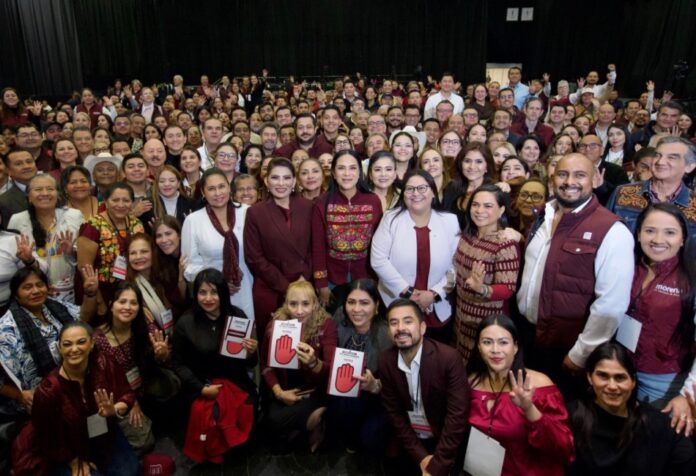Grupo numeroso de integrantes de Morena posa frente a cortinas negras; varias personas levantan las manos y muestran acreditaciones del partido.