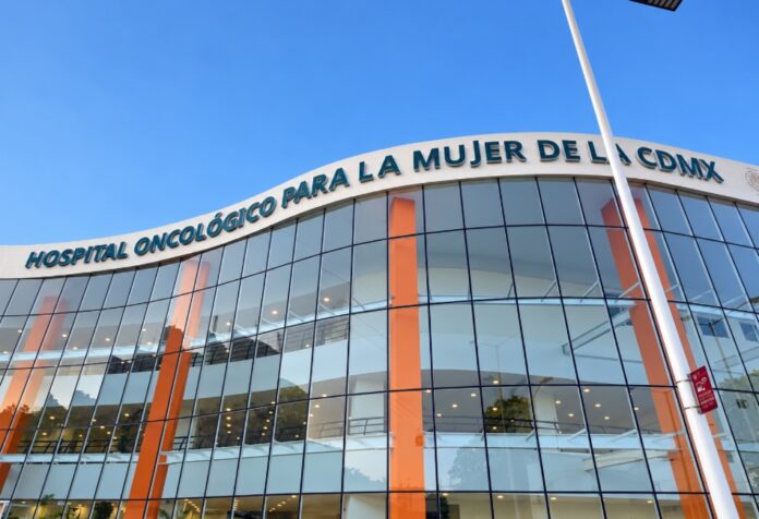 Fachada de cristal del Hospital Oncológico para la Mujer de la Ciudad de México, con columnas anaranjadas y su nombre sobre la entrada.