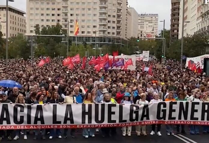 Multitud en una manifestación por la vivienda, con banderas y una pancarta que llama a la huelga general.