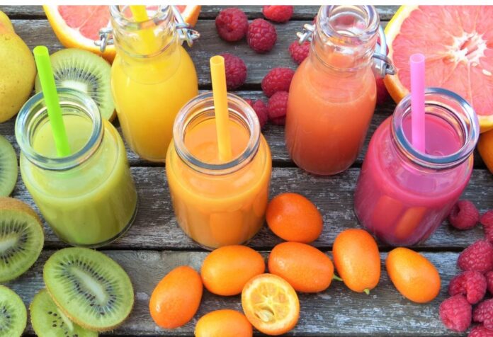 Jugos de frutas Vaso de jugo de naranja junto a un plato con naranja, toronja, kiwi y frambuesas sobre una mesa de madera.
