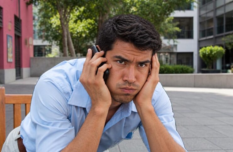 Senado aprueba multas millonarias contra las llamadas publicitarias insistentes Hombre sentado en una banca sostiene un teléfono junto al oído y se apoya la cabeza en la otra mano, con expresión de molestia.