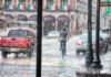 Llueve a cántaros, pero el agua sigue faltando Un ciclista y varios vehículos circulan bajo una lluvia intensa en una calle con edificios antiguos y banderas mexicanas.