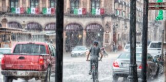 Llueve a cántaros, pero el agua sigue faltando Un ciclista y varios vehículos circulan bajo una lluvia intensa en una calle con edificios antiguos y banderas mexicanas.