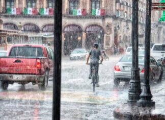 Llueve a cántaros, pero el agua sigue faltando Un ciclista y varios vehículos circulan bajo una lluvia intensa en una calle con edificios antiguos y banderas mexicanas.