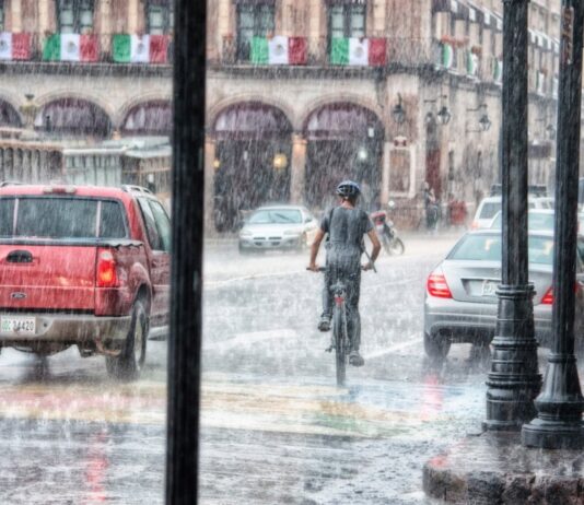 Llueve a cántaros, pero el agua sigue faltando Un ciclista y varios vehículos circulan bajo una lluvia intensa en una calle con edificios antiguos y banderas mexicanas.