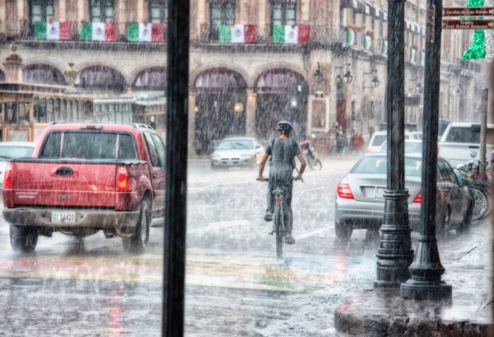 Un ciclista y varios vehículos circulan bajo una lluvia intensa en una calle con edificios antiguos y banderas mexicanas.