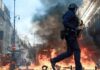 Francia: las protestas por educación y costo de vida llevan al gobierno a imponer toques de queda Policía con casco y escudo camina frente a llamas y una columna de humo entre edificios.