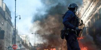 Francia: las protestas por educación y costo de vida llevan al gobierno a imponer toques de queda Policía con casco y escudo camina frente a llamas y una columna de humo entre edificios.
