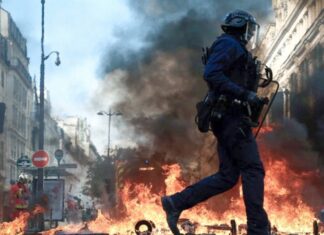 Francia: las protestas por educación y costo de vida llevan al gobierno a imponer toques de queda Policía con casco y escudo camina frente a llamas y una columna de humo entre edificios.