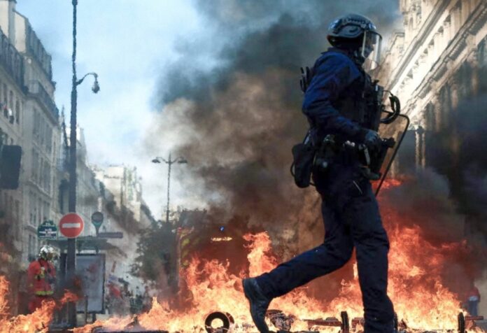 Protestas en Francia Policía con casco y escudo camina frente a llamas y una columna de humo entre edificios.