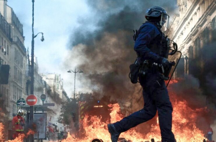 Francia: las protestas por educación y costo de vida llevan al gobierno a imponer toques de queda Policía con casco y escudo camina frente a llamas y una columna de humo entre edificios.