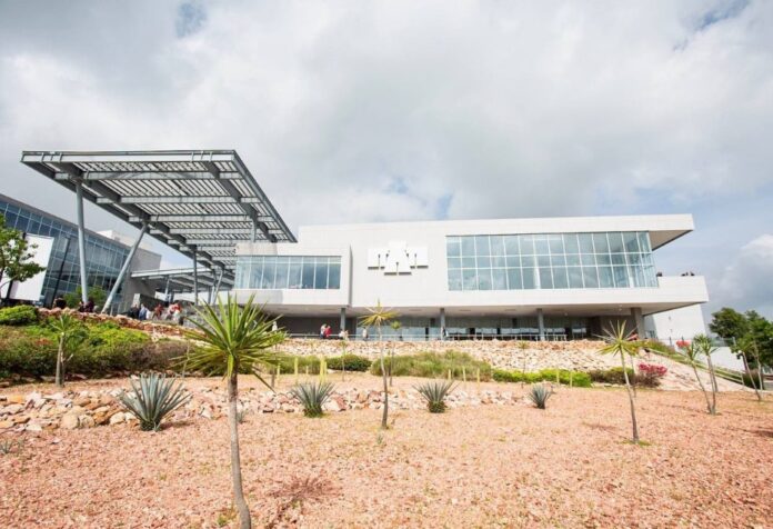 Edificio universitario de fachada blanca y grandes ventanales, con una estructura metálica y jardines de plantas de zonas áridas.