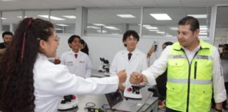 La Mixteca poblana amplía sus oportunidades para estudiar medicina y enfermería Una estudiante con bata blanca saluda a un funcionario del Gobierno de Puebla en un laboratorio de la Universidad de la Salud, mientras otros estudiantes observan. Sobre las mesas hay microscopios y equipo de laboratorio.