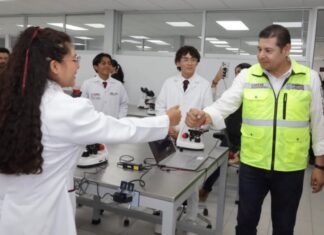 La Mixteca poblana amplía sus oportunidades para estudiar medicina y enfermería Una estudiante con bata blanca saluda a un funcionario del Gobierno de Puebla en un laboratorio de la Universidad de la Salud, mientras otros estudiantes observan. Sobre las mesas hay microscopios y equipo de laboratorio.