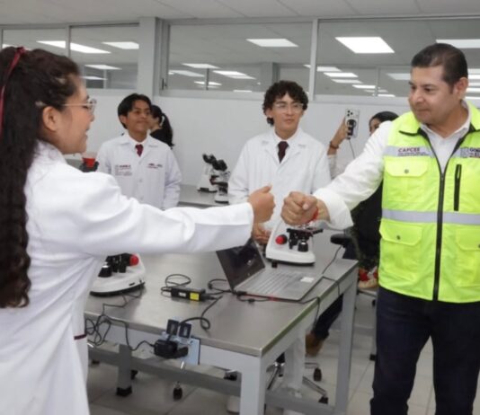 La Mixteca poblana amplía sus oportunidades para estudiar medicina y enfermería Una estudiante con bata blanca saluda a un funcionario del Gobierno de Puebla en un laboratorio de la Universidad de la Salud, mientras otros estudiantes observan. Sobre las mesas hay microscopios y equipo de laboratorio.