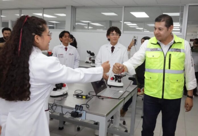 Universidad de la Salud en Puebla Una estudiante con bata blanca saluda a un funcionario del Gobierno de Puebla en un laboratorio de la Universidad de la Salud, mientras otros estudiantes observan. Sobre las mesas hay microscopios y equipo de laboratorio.